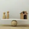 Wooden people figures and a small house balanced on a seesaw, symbolizing the delicate balance and control needed to manage family assets and secure family property